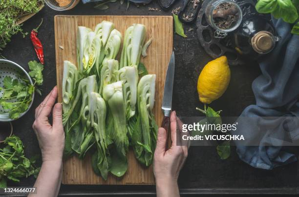 human hands holding wooden cutting board with sliced pak choi and kitchen knife on dark kitchen table with lemon, herbs and dish towel - chinese cabbage stock pictures, royalty-free photos & images