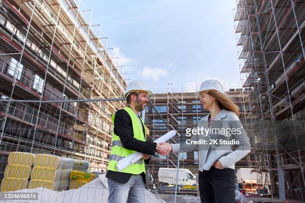 an architect or civil engineer with planning documents meets with a customer or colleague on the large construction site and both of them wear white protective helmets. - civilperson stock pictures, royalty-free photos & images