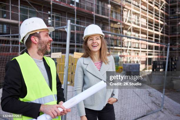 ein architekt oder bauingenieur mit planungsunterlagen trifft sich mit einem kunden oder kollegen auf der großbaustelle und beide tragen weiße schutzhelme. - bauingenieur stock-fotos und bilder