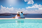 A elegant couple in summer clothes sits by the pool and enjoys the view to the mediterranean sea