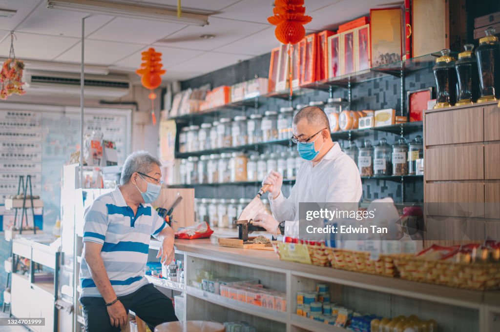 Asian chinese male pharmacist cutting ginseng chinese herbs medicine for his senior patient at traditional chinese herbal medicine shop