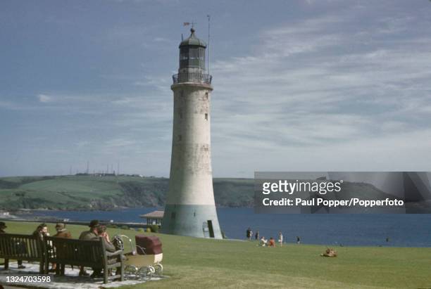 Visitors sit on benches in front of Smeaton's Tower at Plymouth Hoe with the Mount Batten peninsula, Batten Bay and Plymouth Sound beyond, in Devon,...