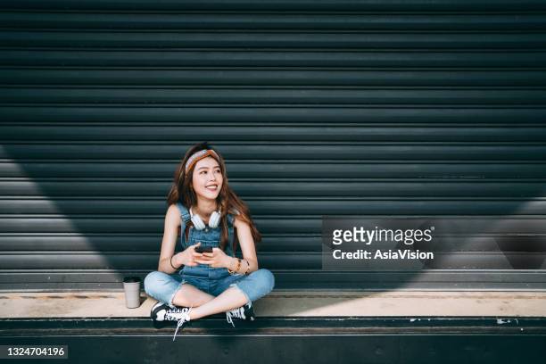 smiling asian female university student wearing headphones around her neck, looking up while text messaging on smartphone, sitting in front of roller shutter in school campus against sunlight. teenage lifestyle and technology - beautiful asian student stockfoto's en -beelden