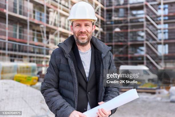 un arquitecto o ingeniero civil se para con documentos de planificación enrollados frente a un edificio de apartamentos con andamios y lleva un casco blanco. - civil engineering fotografías e imágenes de stock