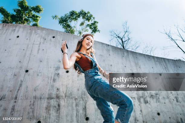 retrato de ángulo bajo de una joven asiática despreocupada sosteniendo un teléfono inteligente, bailando mientras escucha música en auriculares al aire libre contra la pared de concreto y la luz del sol. música y estilo de vida - action laptop fotografías e imágenes de stock