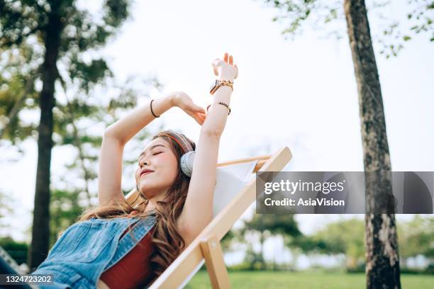 beautiful, carefree young asian woman with her eyes closed relaxing on deck chair in park, listening to music with headphones and hands raised. enjoying summer days outdoors. music, teenage lifestyle and technology - girl headphones imagens e fotografias de stock