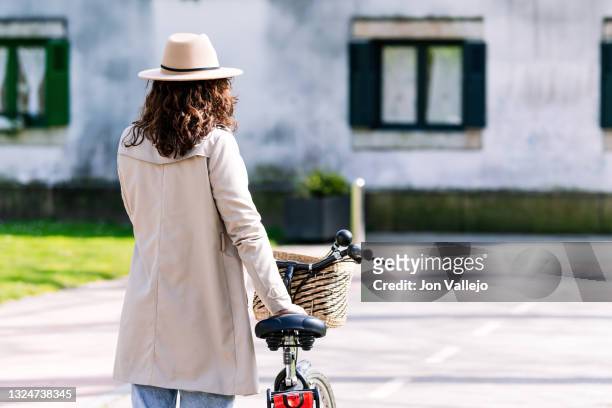 foto de espaldas de una mujer que tiene el pelo rizado. ella va caminando al lado de su bicicleta. lleva puesto una gabardina de color beige y un sombrero del mismo color. - mujer de espaldas foto e immagini stock