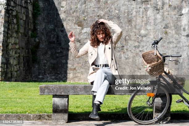 mujer guapa esta sentada en un banco de hormigon mientras se esta arreglando su pelo rizado. en el banco hay un bolso beige que va a juego con su gabardina y al lado del banco esta su bicicleta negra. - mujer negra stock pictures, royalty-free photos & images