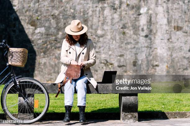 una mujer que esta sentada junto a su bicicleta sobre un banco de hormigon, busca algo dentro de su pequeño bolso de cuero. la mujer tiene a su izquierda sobre el banco, un ordenador portatil pequeño y lleva puesto una gabardina de color beige y un sombr - ordenador stock-fotos und bilder