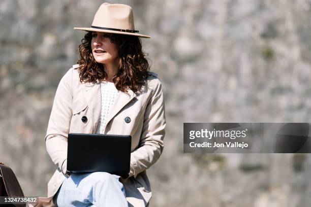 mujer joven con el pelo rizado, esta sentada con un ordenador portatil pequeño sobre sus piernas. la mujer lleva puesto un sombrero de color beige y una gabardina del mismo color. - ordenador stock-fotos und bilder