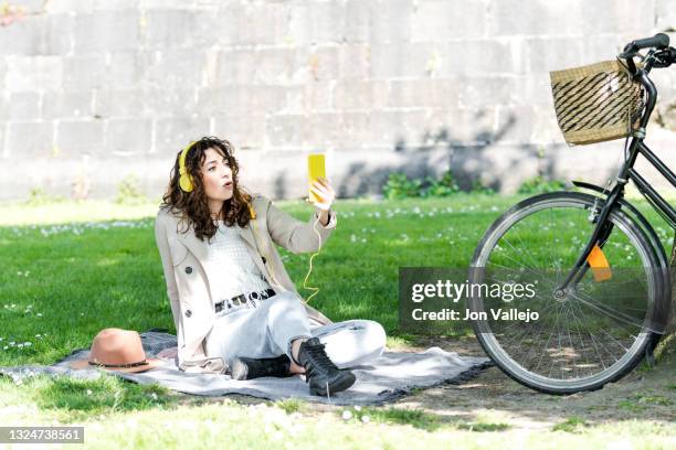 chica muy guapa de pelo rizado esta con cara de sorprendida mientras esta mirando su movil con carcasa amarilla. la mujer lleva puestos unos auriculares amarillos con cable y esta sentada encima de una manta en la hierba junto a su bicicleta que esta apoy - chica movil stockfoto's en -beelden