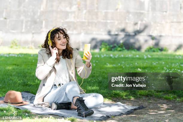 mujer muy guapa de pelo rizado esta sonriendo sujetando con la mano sus auriculares con cable amarillos. la mujer esta haciendo una video llamada con el movil con carcasa amarilla que tiene en la otra mano y esta sentada sobre una manta encima de la hierb - movil mano stock-fotos und bilder