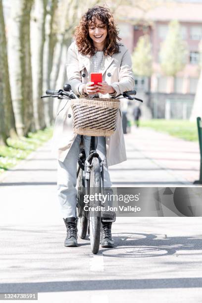 mujer atractiva esta sonriendo mientras esta mirando su movil con carcasa roja mientras esta de pie con su bicicleta sobre el carril bici. la bicicleta tiene una cesta de mimbre en la parte delantera y la mujer lleva puesto una gabardina de color beige. - bici mujer ストックフォトと画像