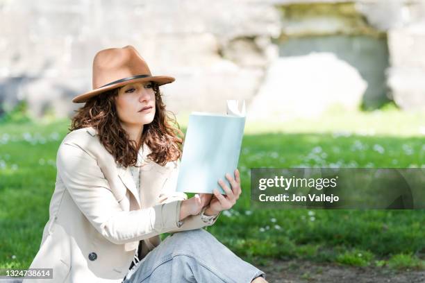 una chica joven muy guapa de pelo rizado esta pensativa mientras sujeta un libro con las mano. la mujer esta sentada en la hierba y lleva puesto una gabardina de color beige y un sombrero de color marron. - chica pensativa stockfoto's en -beelden