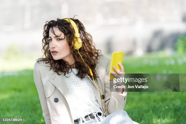 chica muy guapa de pelo rizado esta mirando hacia un lado con su movil con carcasa amarilla en la mano. la mujer lleva puestos unos auriculares amarillos con cable y esta sentada en la hierba. - chica movil stockfoto's en -beelden