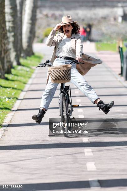 mujer atractiva de pelo rizado viene con cara de asustada con la boca abierta mientras sujeta con una mano su sombrero de color beige que va a juego con su gabardina. la mujer va encima de su bicicleta por el carril bici con las piernas abiertas fuera de - bici mujer stock pictures, royalty-free photos & images