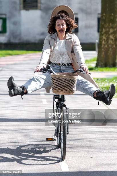 mujer atractiva de pelo rizado viene riendose mientras se le esta cayendo su sombrero de color beige y este va a juego con su gabardina. la mujer va encima de su bicicleta por el carril bici mientras va con las piernas abiertas fuera de los pedales. - bici mujer stock pictures, royalty-free photos & images