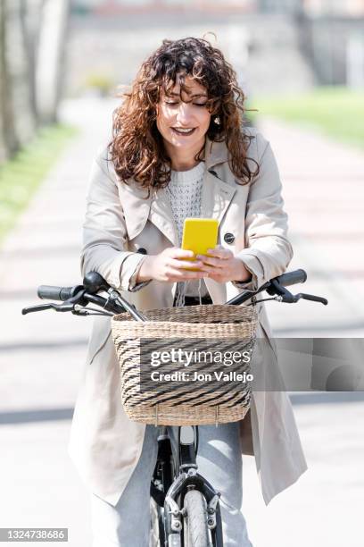 primer plano de una mujer muy guapa de pelo rizado que sonrie mientras esta mirando su movil con carcasa amarilla. la mujer esta de pie con su bicicleta y esta tiene una cesta de mimbre en la parte delantera. esta en el carril bici. - bici mujer stock pictures, royalty-free photos & images