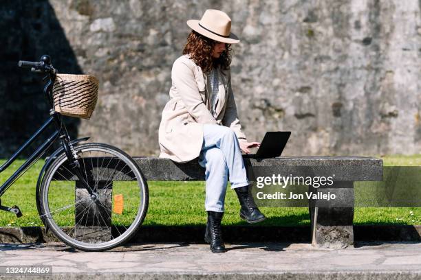 una chica joven muy guapa con el pelo rizado esta sentada en un banco mientras trabaja con un pequeño ordenador portatil negro. la mujer tiene su bicicleta a su lado y lleva puesto una gabardina de color beige y un sombrero del mismo color. - ordenador stock-fotos und bilder