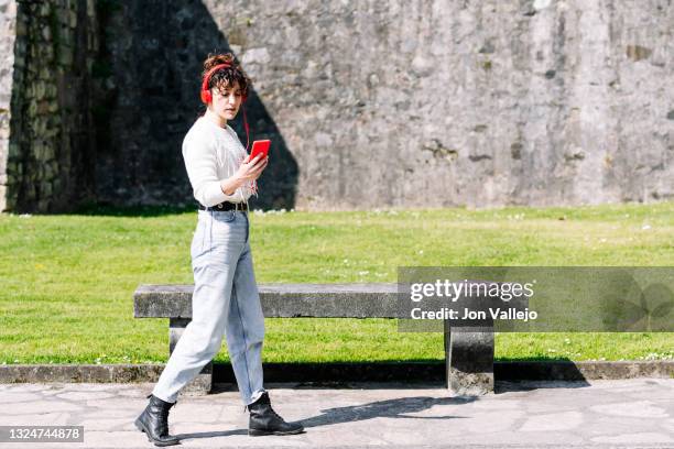 mujer atractiva camina mientras mira su movil con funda de color rojo. el movil esta conectado a sus auriculares de color rojo por mediacion de un cable. - mediacion photos et images de collection