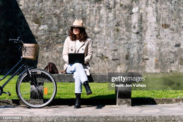 mujer atractiva con el pelo rizado, esta sentada en un banco de hormigon junto a su bicicleta mientras trabaja con un pequeño ordenador portatil. la mujer lleva puesto un sombrero beige y una gabardina del mismo color. - ordenador stock-fotos und bilder