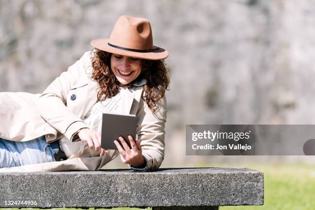 una mujer se esta riendo mientras mira una tableta que tiene en sus manos. la mujer de pelo rizado esta tumbada de lado sobre un banco de hormigon y lleva puesto una gabardina color beige y un sombrero de color marron. - tableta digital stock pictures, royalty-free photos & images