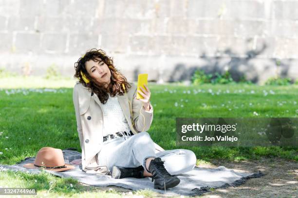 mujer muy guapa de pelo rizado esta con la mirada perdida mientras sujeta su movil con carcasa amarilla con la mano. la chica esta sentada encima de una manta encima de la hierba. - chica movil stockfoto's en -beelden