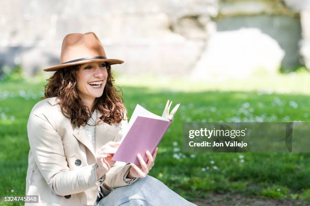 una chica joven muy guapa de pelo rizado esta sentada en la hierba mientras sonrie. la mujer esta con un libro en las manos y lleva puesto un sombrero marron. - mujer sentada stock pictures, royalty-free photos & images