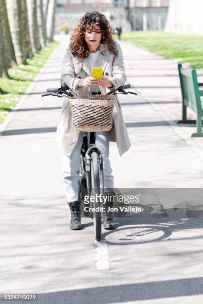 una mujer atractiva de pelo rizado esta parada de pie con su bicicleta en el carril bici esta, tiene una cesta de mimbre en la parte delantera. la mujer esta mirando su movil con carcasa amarilla y lleva puesto una gabardina de color beige. - bici mujer ストックフォトと画像