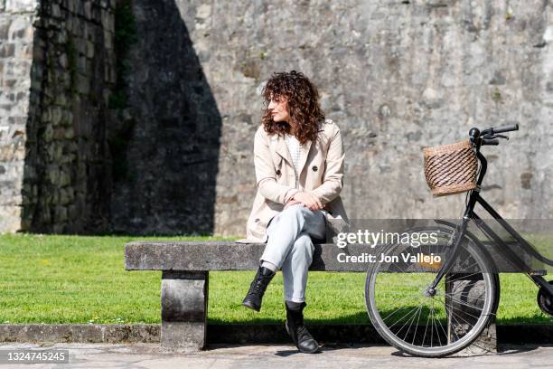 mujer muy guapa esta sentada en un banco de hormigon junto a su bicicleta negra. la mujer esta mirando hacia su derecha y lleva puesto una gabardina de color beige. - mujer negra stock-fotos und bilder