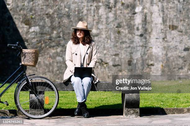 mujer muy guapa con pelo de rizos y un sombrero color beige esta sentada en un banco de hormigon junto a su bicicleta. la mujer esta trabajando con un ordenador portatil pequeño. - mujer sentada stock-fotos und bilder