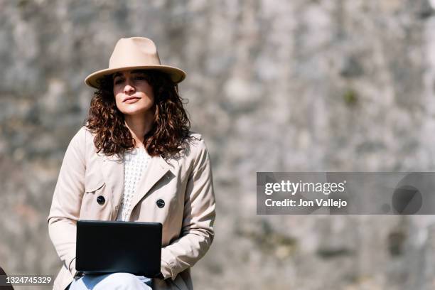 mujer atractiva con el pelo rizado esta pensativa mientras esta trabajando con un pequeño ordenador portatil. la mujer lleva puesto una gabardina de color beige y un sombrero del mismo color. - pensativa stock-fotos und bilder