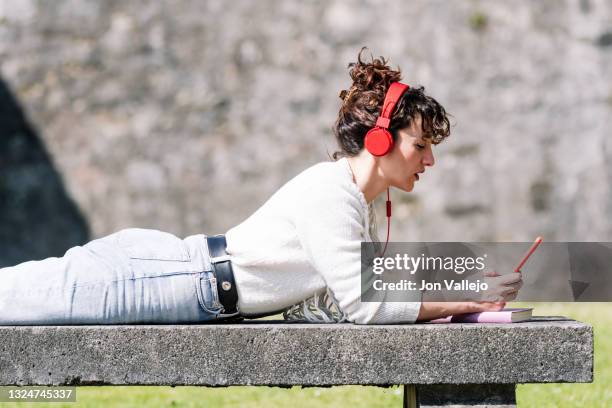 mujer con el pelo rizado mirando su movil de carcasa roja mientras esta tumbada boca abajo en un banco de hormigon. la mujer tiene unos auriculares con cable de color rojo conectados a su movil. - pelo mujer stock pictures, royalty-free photos & images