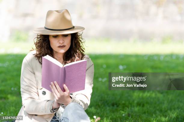primer plano de una mujer muy atractiva de pelo rizado que esta leyendo un libro sentada en la hierba. la mujer lleva puesto un sombrero de color beige y una gabardina a juego. - mujer sentada stock pictures, royalty-free photos & images
