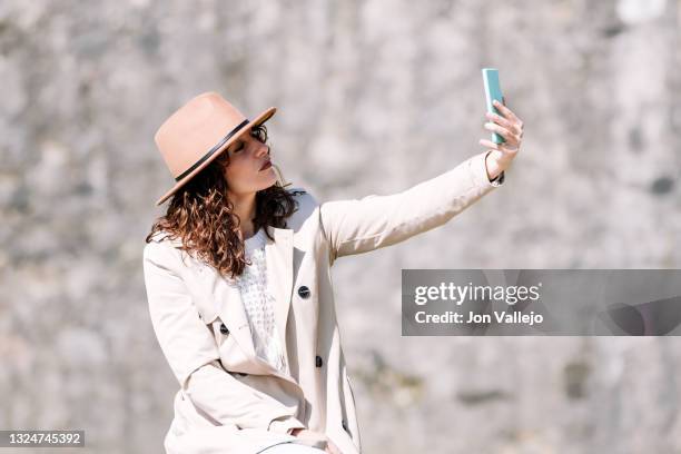 mujer atractiva de pelo rizado se esta haciendo una autofoto con su movil de carcasa azul. la mujer lleva puesto un sombrero de color marron y una gabardina de color beige. - pelo mujer stock pictures, royalty-free photos & images