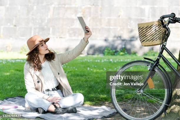 chica muy guapa de pelo rizado esta sonriendo mientras se esta haciendo una autofoto con su tablet mientras esta sentada encima de una manta en la hierba junto a su bicicleta que esta apoyada en un arbol. - chica sonriendo stock pictures, royalty-free photos & images