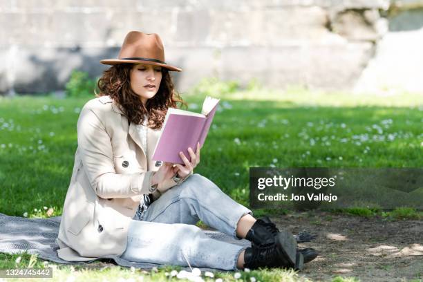 mujer muy guapa esta concentrada mientras lee un libro sentada en una manta en la hierba. la mujer lleva puesto una gabardina de color beige y un sombrero marron. - mujer sentada stock pictures, royalty-free photos & images