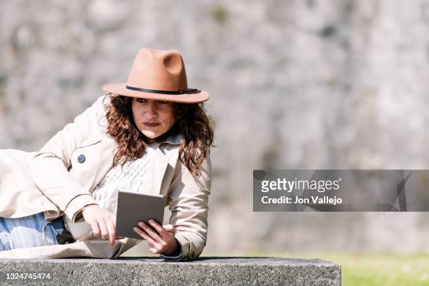 una mujer de pelo rizado tumbada de lado sobre un banco de hormigon. la mujer tiene una tableta en la mano y lleva puesto un sombrero de color marron y una gabardina de color beige. - pelo mujer stock pictures, royalty-free photos & images