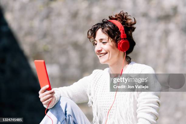 primer plano de una mujer muy guapa que tiene el pelo rizado. la mujer esta sonriendo mientras mira su telefono movil de carcasa roja que tiene en su mano y en el movil esta enchufado un cable de sus auriculares rojos. - primer plano stock-fotos und bilder