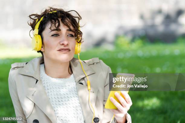 primer plano de una mujer muy atractiva que esta mirando a lo lejos mientras esta sentada en la hierba con su movil de carcasa amarilla en la mano. la mujer esta con unos auriculares con cable amarillos sobre la cabeza. - mujer sentada stock pictures, royalty-free photos & images