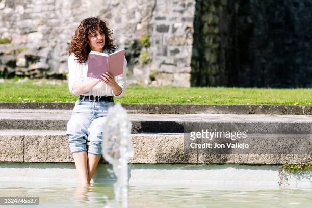 mujer muy guapa de pelo rizado esta sonriendo sentada en el suelo con los pies en una fuente mientras esta leyendo un libro que tiene en las manos. - mujer sentada stock-fotos und bilder