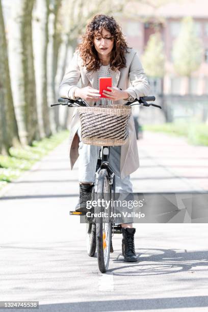 mujer muy guapa de pelo rizado esta sobre su bicicleta parada mirando su movil con carcasa roja. esta en el carril bici. - bici mujer stock pictures, royalty-free photos & images