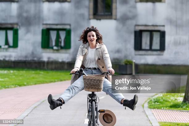 mujer de pelo rizado va con las piernas abiertas sobre su bicicleta por el carril bici. a la mujer se le acababa de caer su sombrero de color beige que llevaba puesto. el sombrero es del mismo color que la gabardina que lleva puesta. - bici mujer ストックフォトと画像