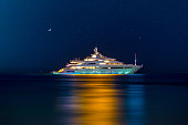 Night view to large illuminated white boat located over horizon, colorful lights coming from yacht reflect on the surface of the the Gulf sea. Shot at blue hour.