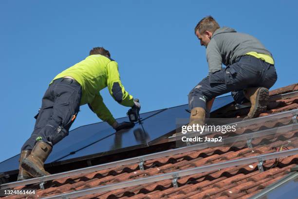 zwei männer auf einem ziegeldach installieren sonnenkollektoren - zonnepanelen stock-fotos und bilder