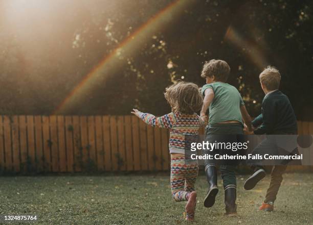 three children hold hands and run across a garden - rainbow kids stock-fotos und bilder