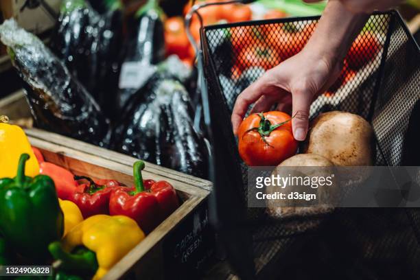cropped shot of young woman grocery shopping for fresh organic produces in a local grocery store, putting a tomato and portobello mushrooms into a shopping basket. green living. healthy eating lifestyle. supporting local business concept - gemüse supermarkt stock-fotos und bilder