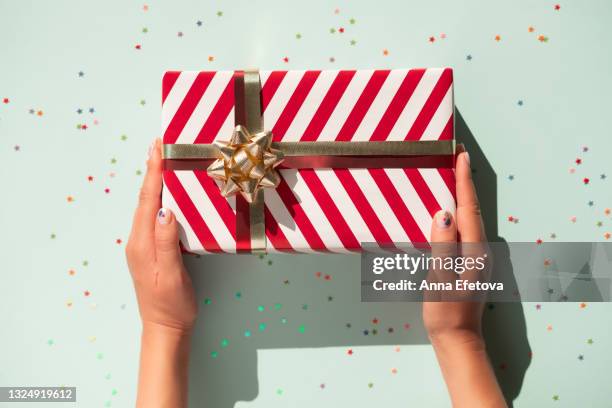 hands are holding gift box with white-red striped pattern and glittering gold bow on pastel light blue background with many star shaped confetti. flat lay style - star confetti white background stockfoto's en -beelden