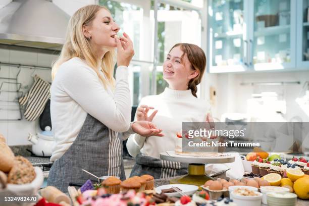 mother and teenage girl daughter baking a cake together in their kitchen. mother tasting a fruitcake is making by her daughter. - woman baking stock-fotos und bilder