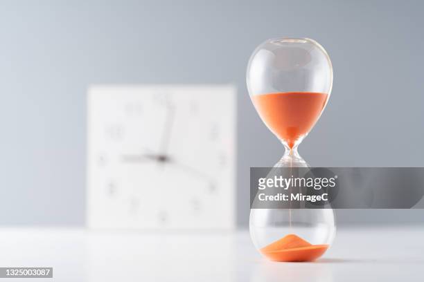 orange colored sand hourglass in front of clock - stechuhr stock-fotos und bilder